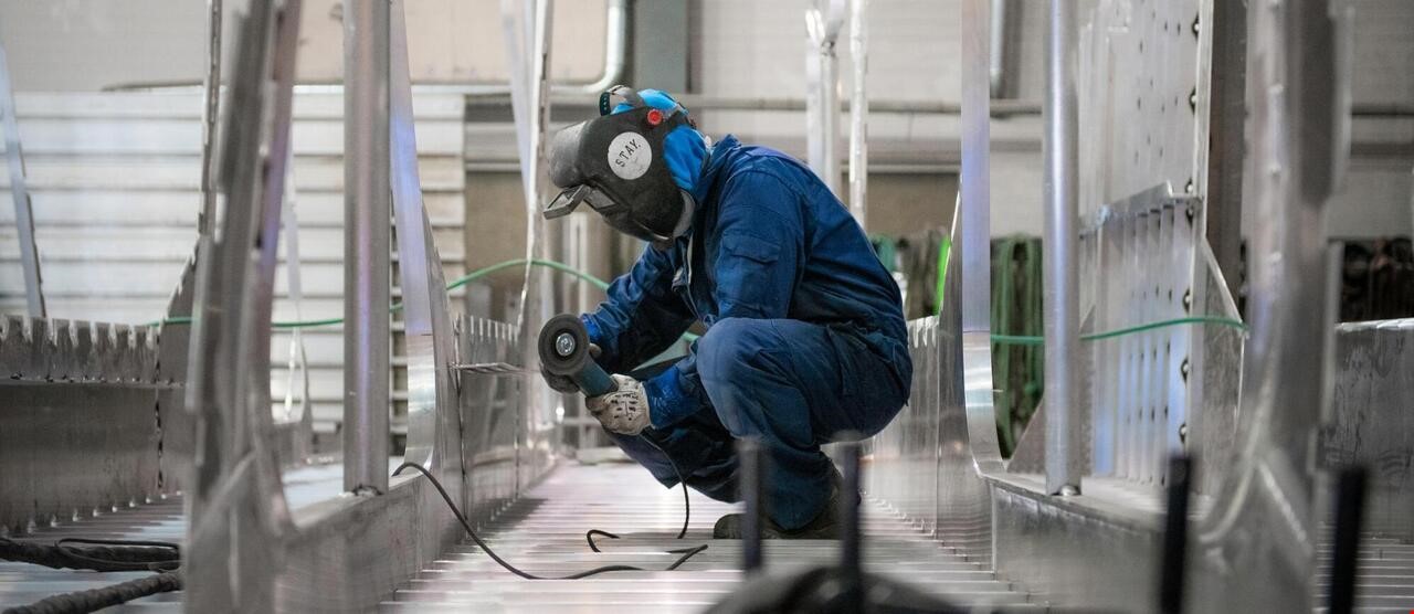 Arbeider iført verneutstyr, sitter på hælene med en elektrisk sag i hendene. Rommet er fylt av diverse aluminiumsplater.